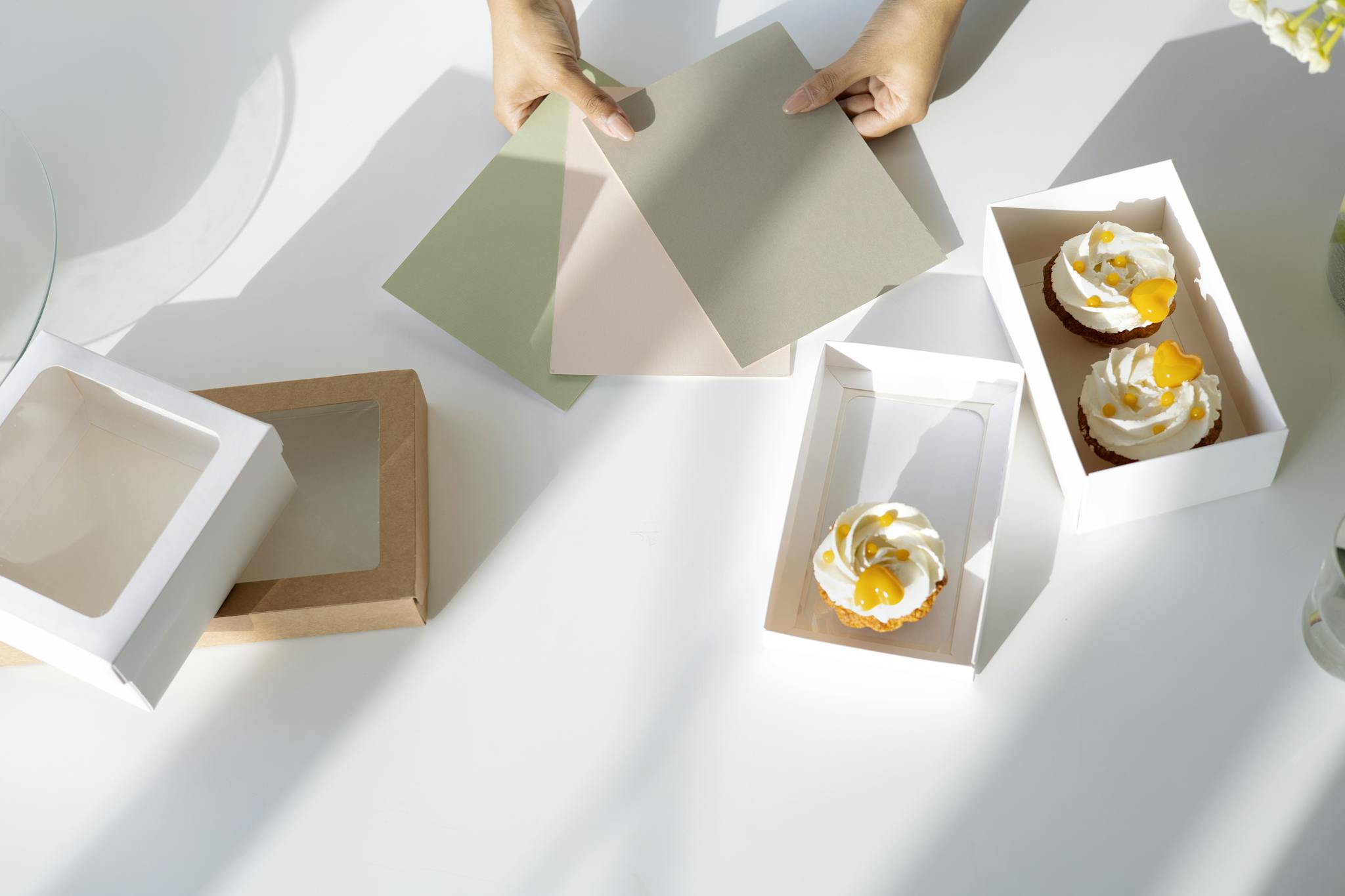 Hands holding color swatches over cupcakes and packaging in a bright setting.