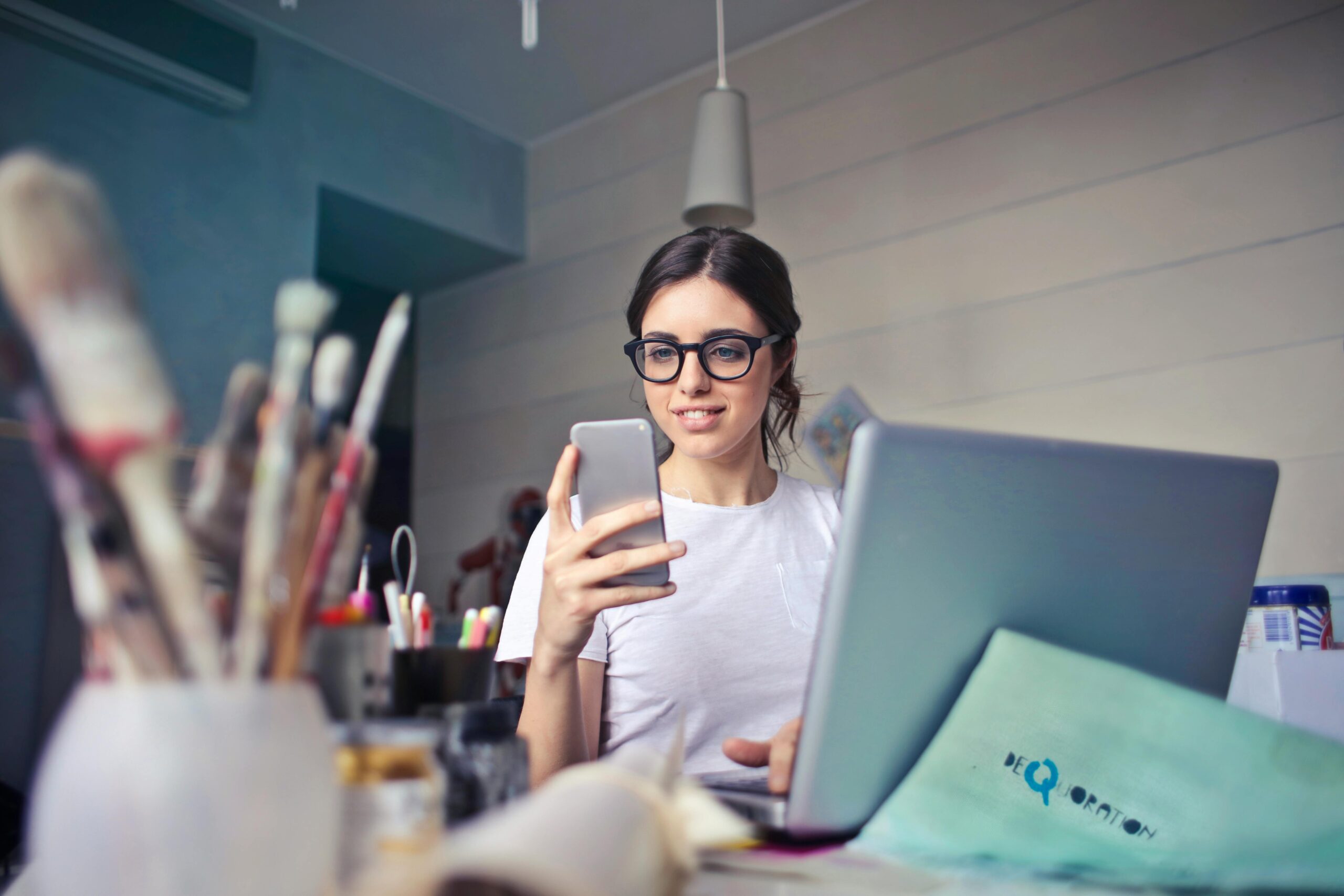 A woman using her phone at a desk, surrounded by art supplies and a laptop, in a creative workspace.